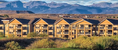 A row of multi-story buildings with balconies, set against a backdrop of mountains and a clear sky.