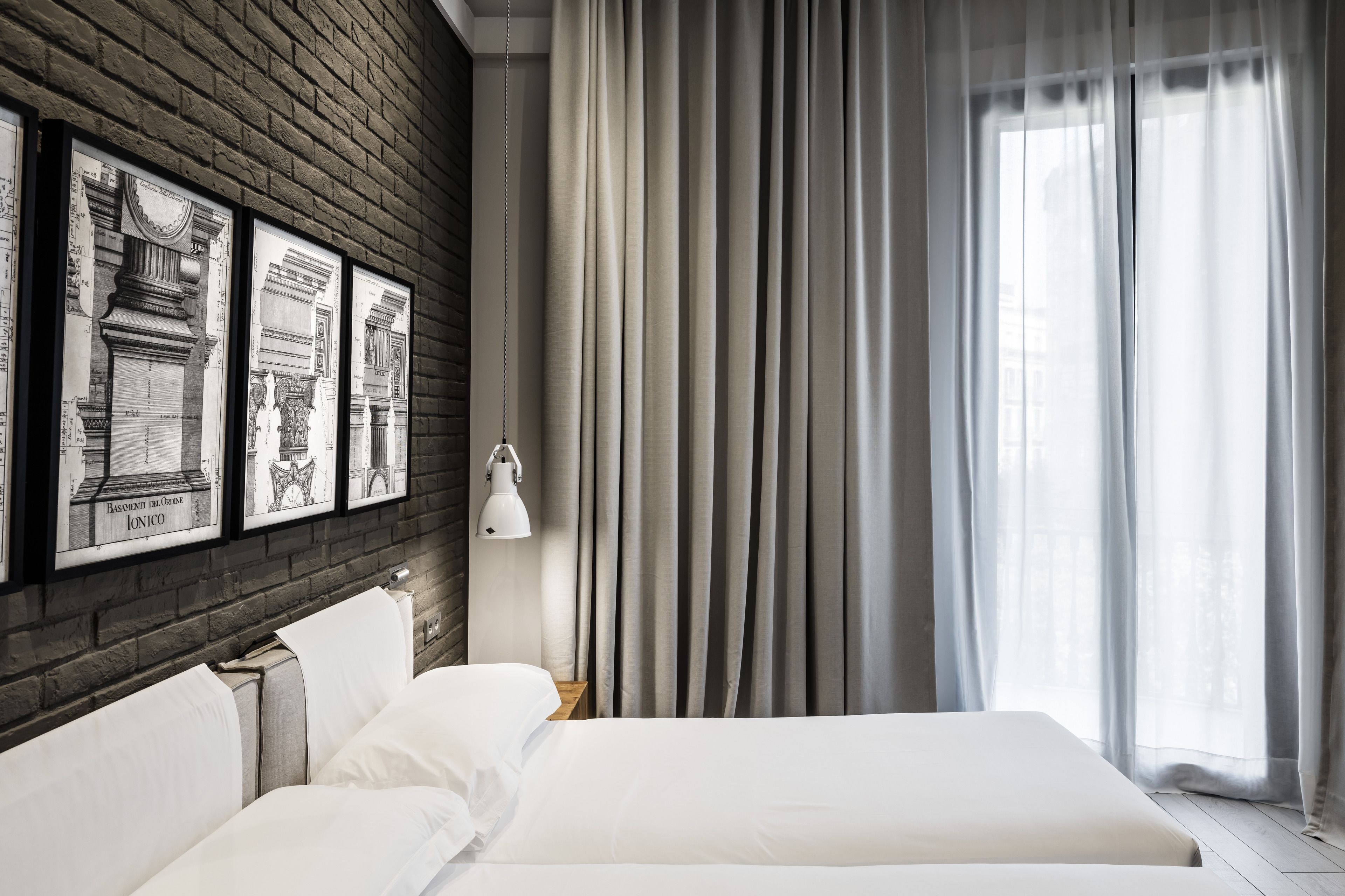 A modern hotel room with a large bed, a nightstand, a lamp, and a window with curtains. There are framed architectural drawings on the wall.