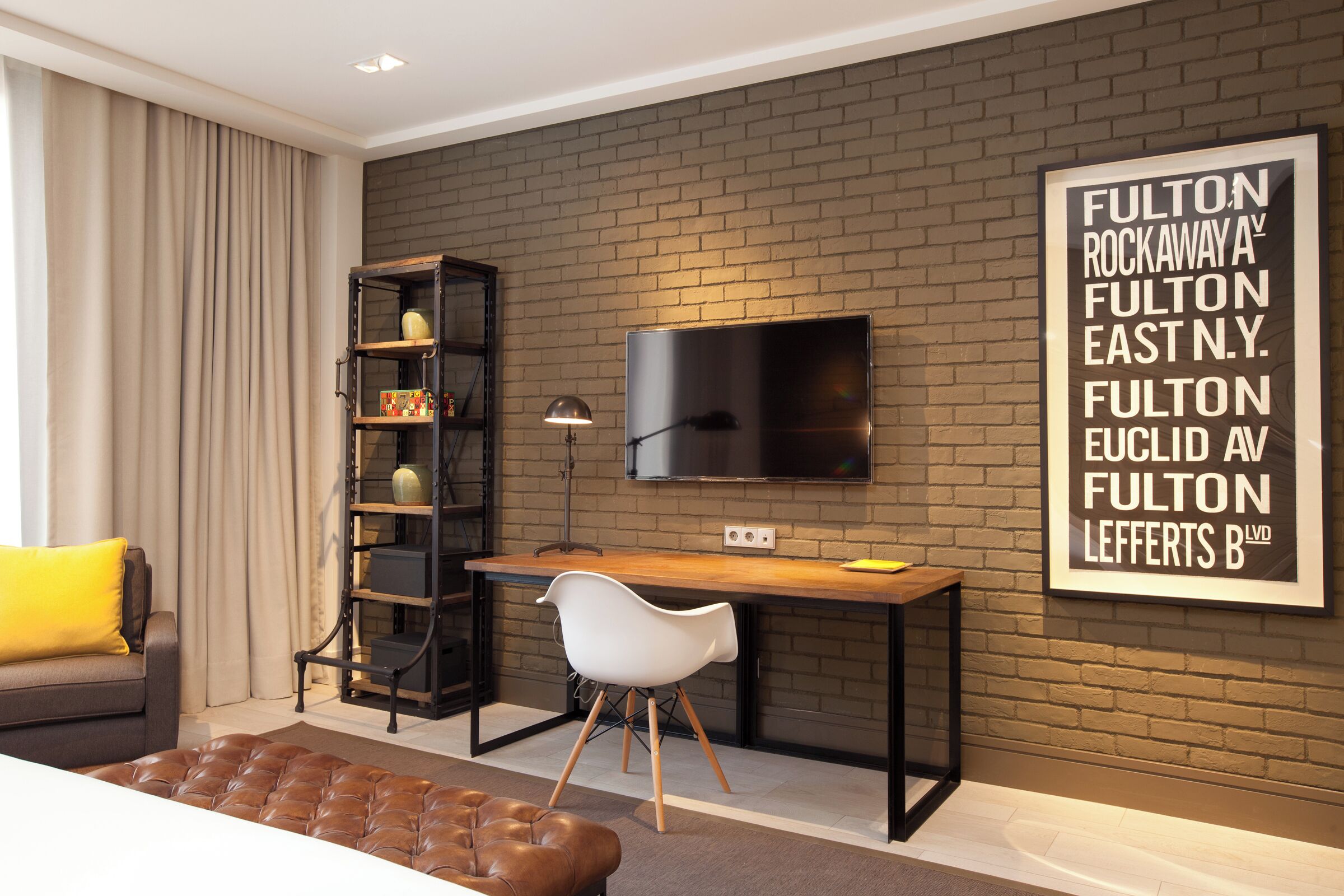Una habitación de hotel moderna con pared de ladrillo, un televisor de pantalla plana montado sobre un escritorio de madera, una silla blanca, un reposapiés de cuero marrón y una estantería.