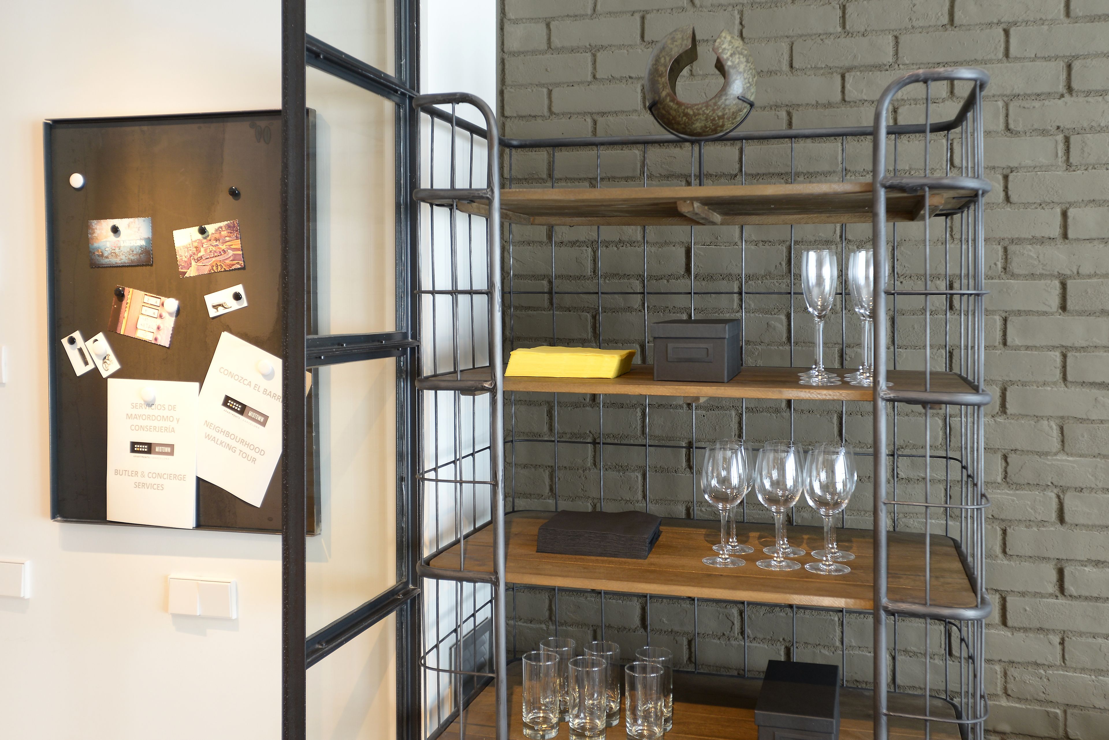 Ein Metallregal mit Weingläsern und einem Korkenzieher auf der obersten Ablage, eine Korktafel mit Notizen auf der linken Seite und eine Backsteinwand im Hintergrund.