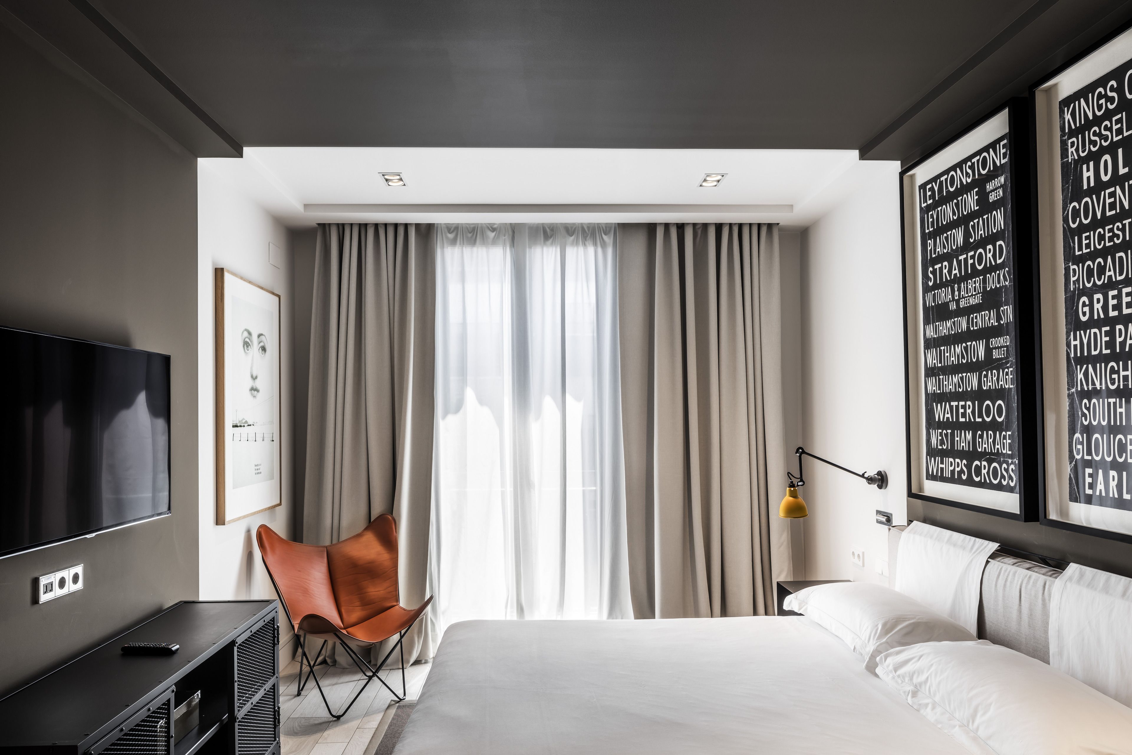 Una habitación de hotel moderna con una cama grande, un televisor de pantalla plana, una silla y un ventanal con cortinas.