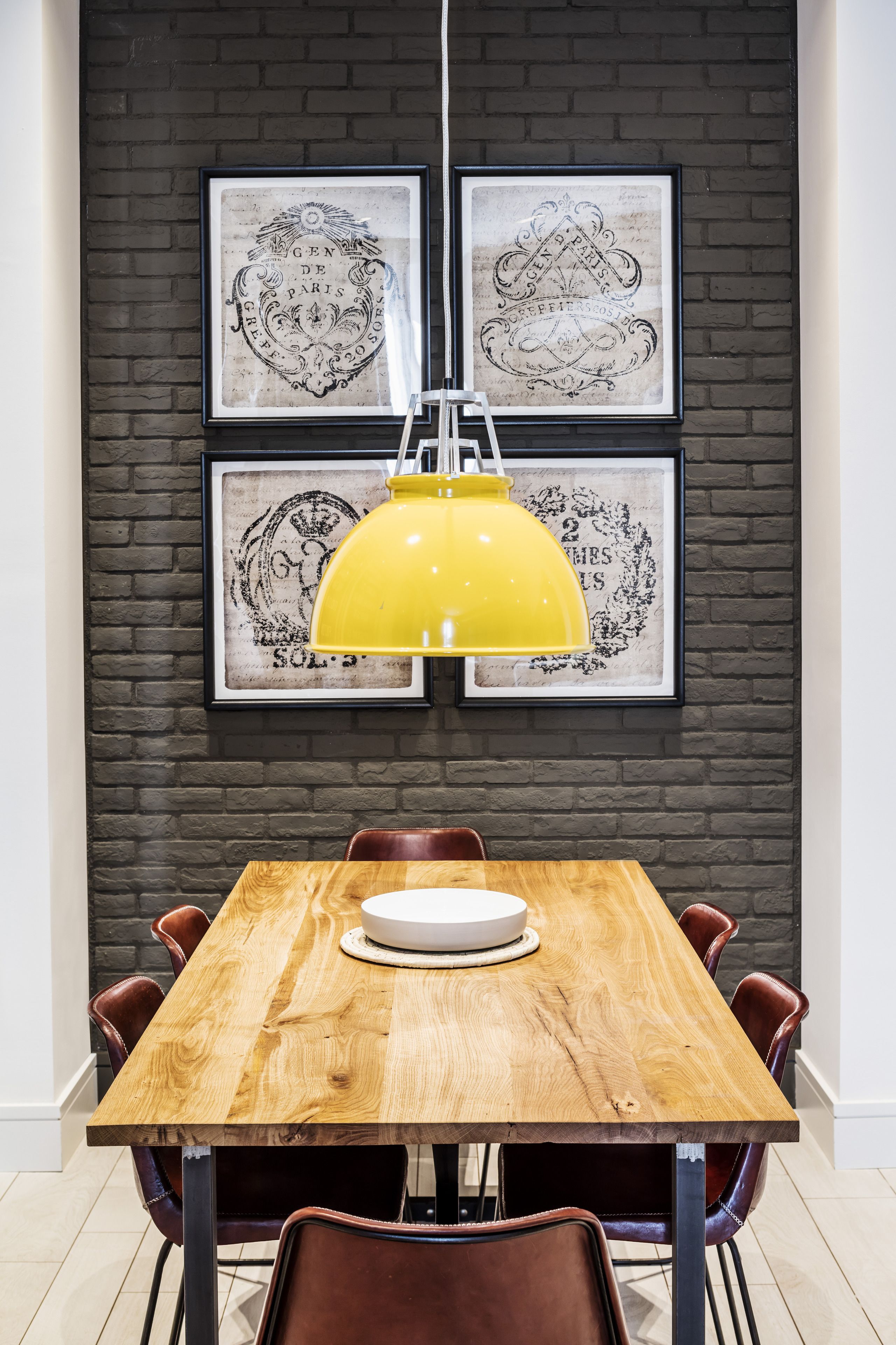 A dining area with a wooden table, four chairs, and a yellow pendant light. There are two framed artworks on the wall above the table.