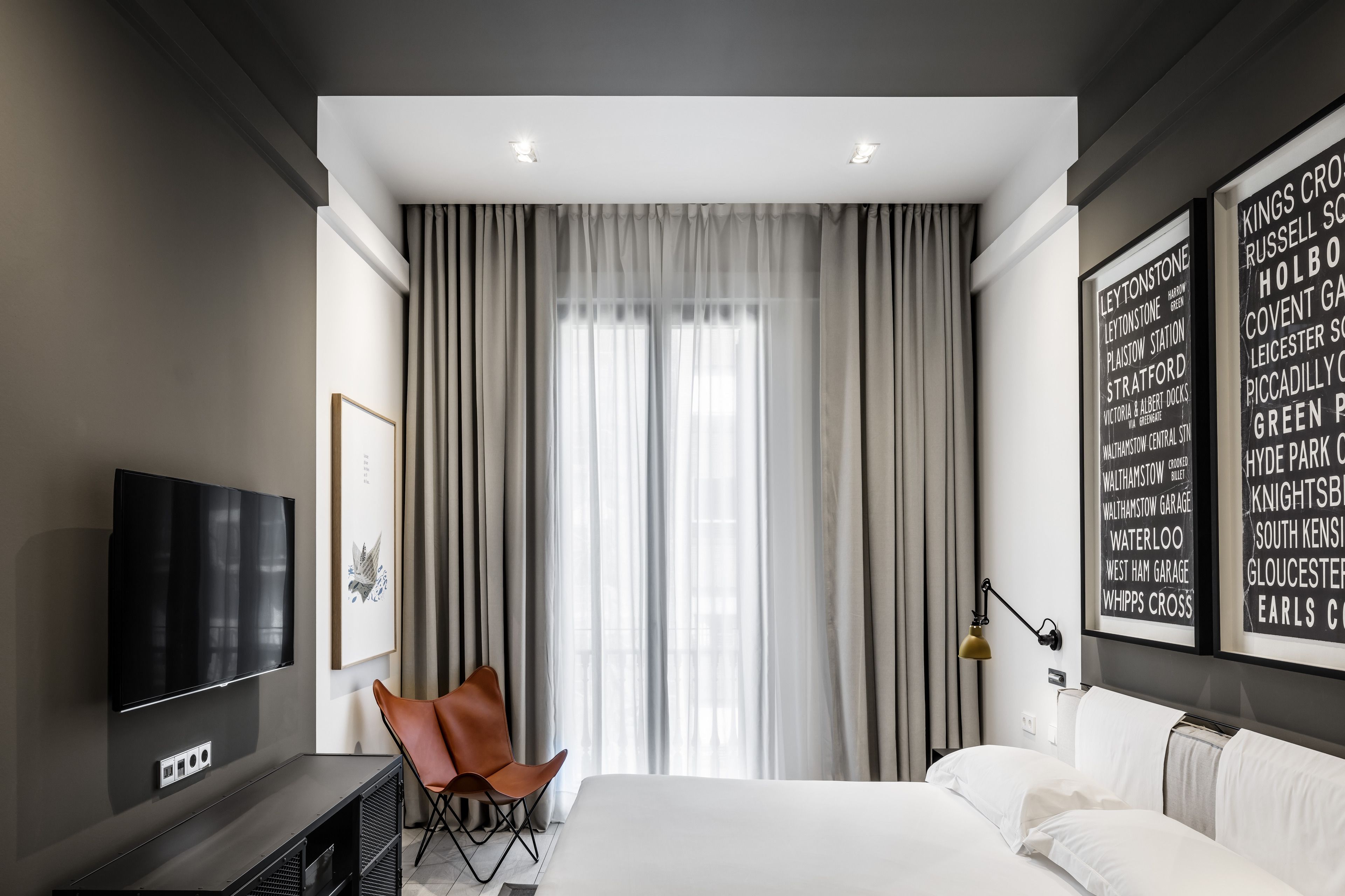 A modern hotel room with a large bed, a flat-screen TV, a seating area with a chair, and a large window with curtains.