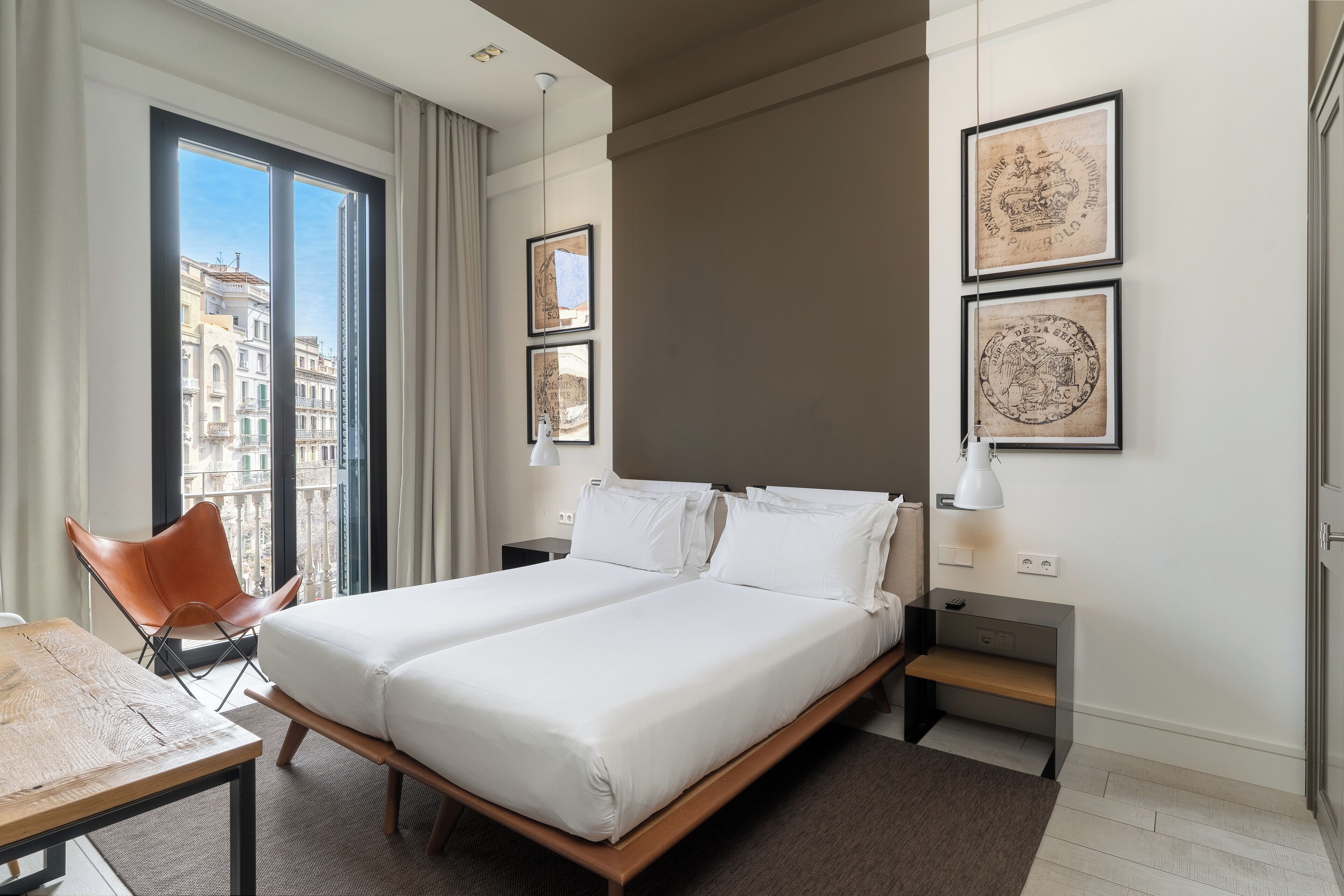Quarto de hotel moderno com uma cama grande, uma escrivaninha e uma cadeira. Há quadros nas paredes e uma vista para edifícios pela janela.