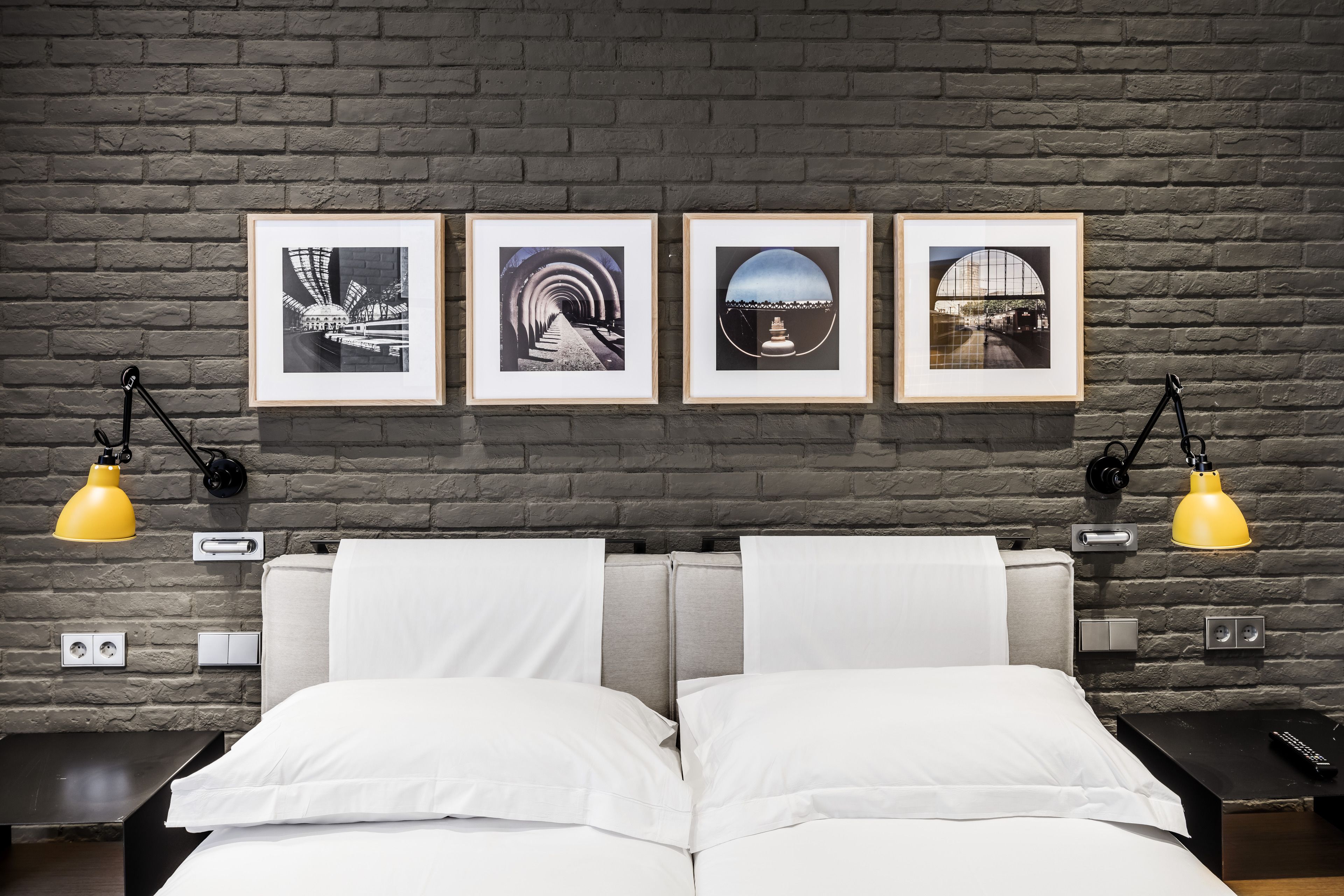 Een hotelkamer met een bakstenen muur, een bed met witte lakens, twee nachtkastjes en drie ingekaderde foto's aan de muur.