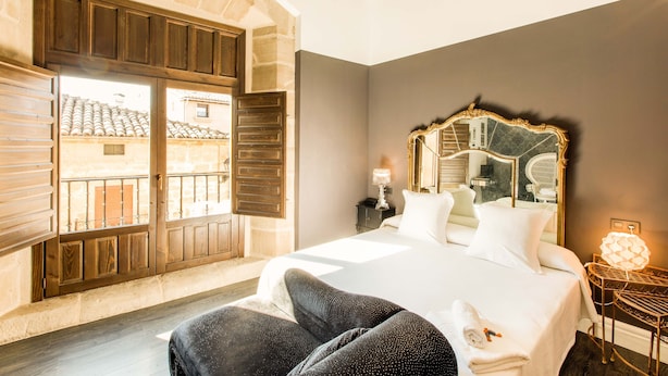 A hotel room with a large bed, a wooden wardrobe, a balcony with a view of the city, and a decorative mirror on the wall. - Teatrisso Hotel Palacio (Cuzcurrita de Rio Tiron)