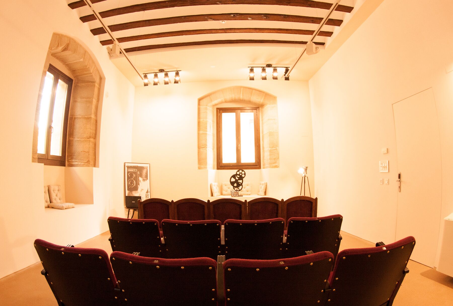 Un teatro con hileras de butacas de terciopelo rojo, un escenario con un piano y un amplio ventanal de arco.
