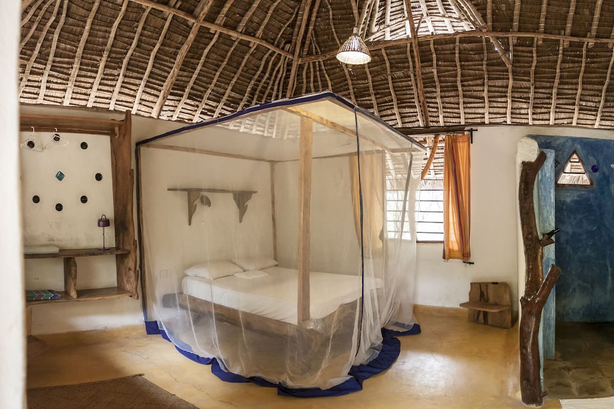 A room with a thatched roof, a bed with a mosquito net, a wooden shelf, a small table, a basin, and a mirror on the wall.