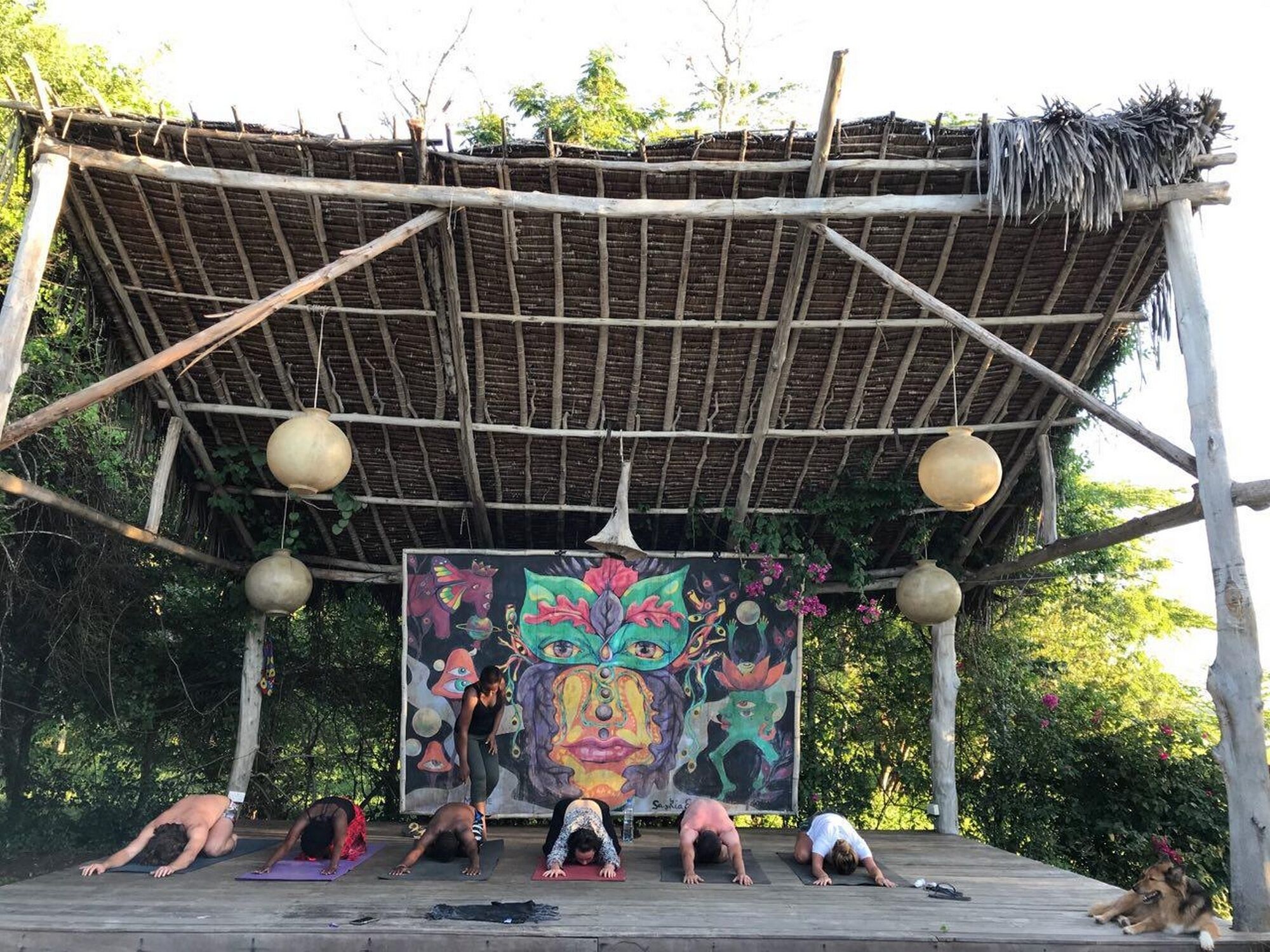 A wooden deck with a thatched roof, a mural, and people practicing yoga.
