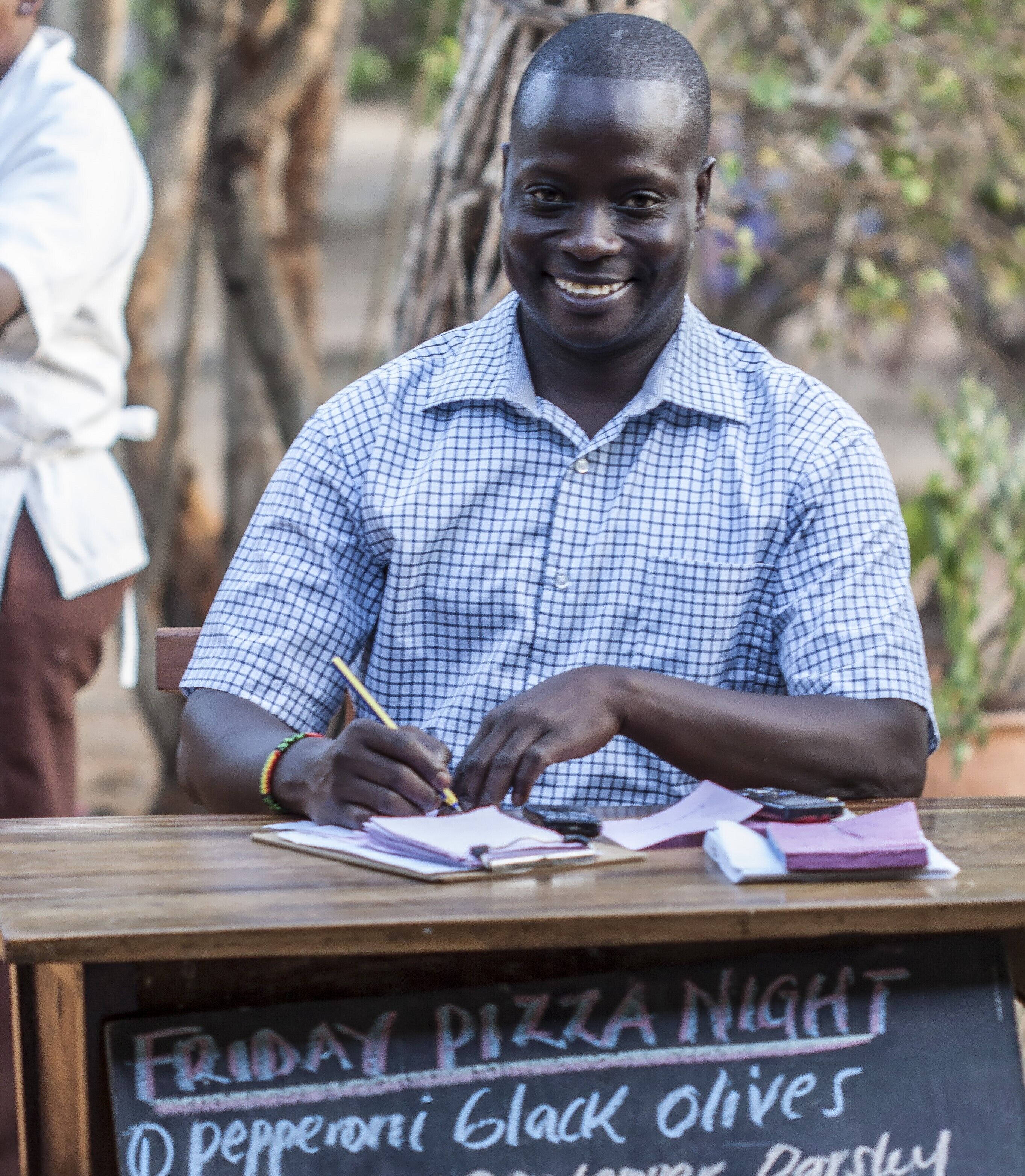A person writing at a wooden table with a chalkboard menu listing items like 'Friday Pizza Night' and 'Pepperoni Black Olives'.