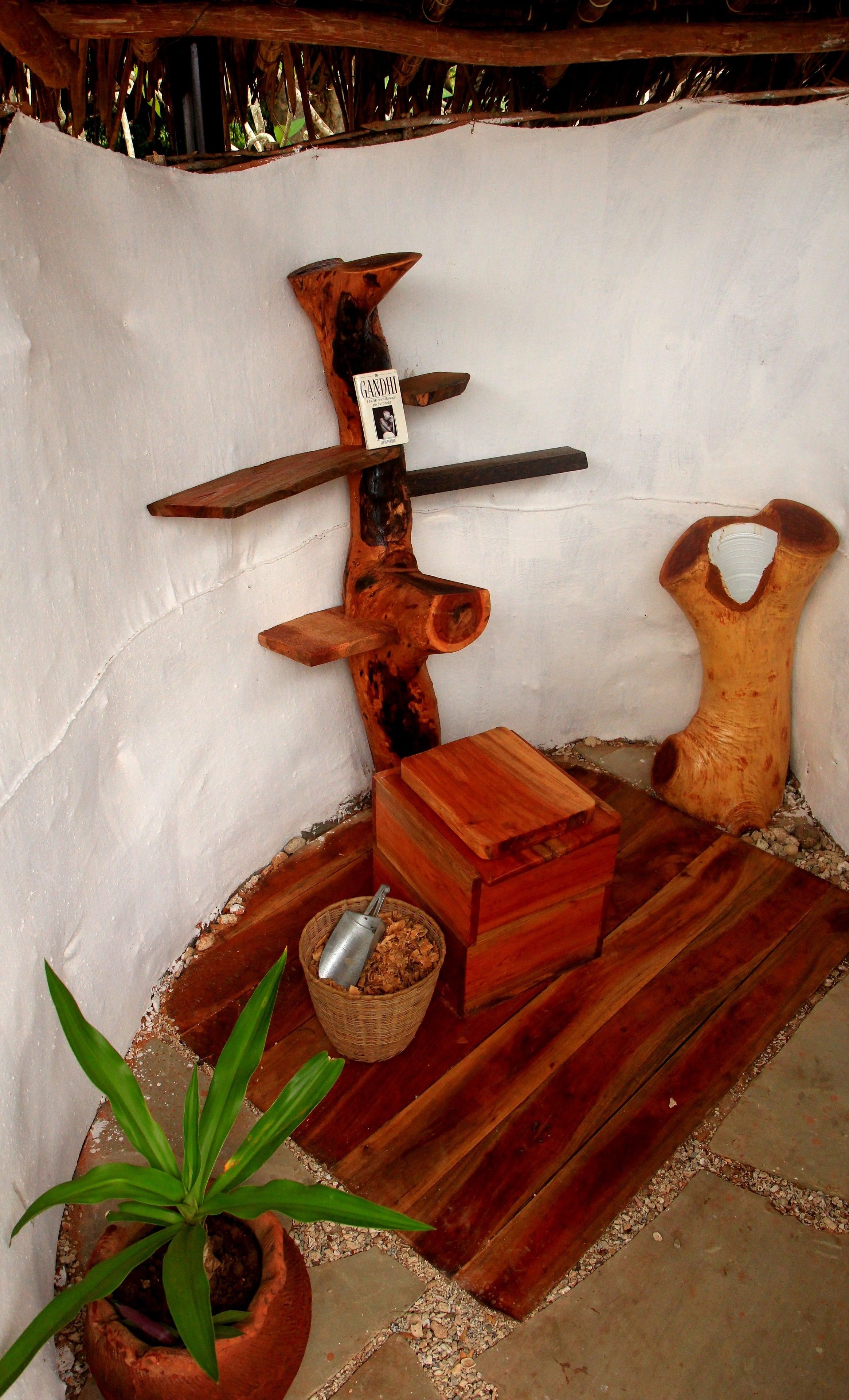 A rustic wooden sculpture with a book titled 'Gandhi' and a metal container on a wooden floor with a potted plant.