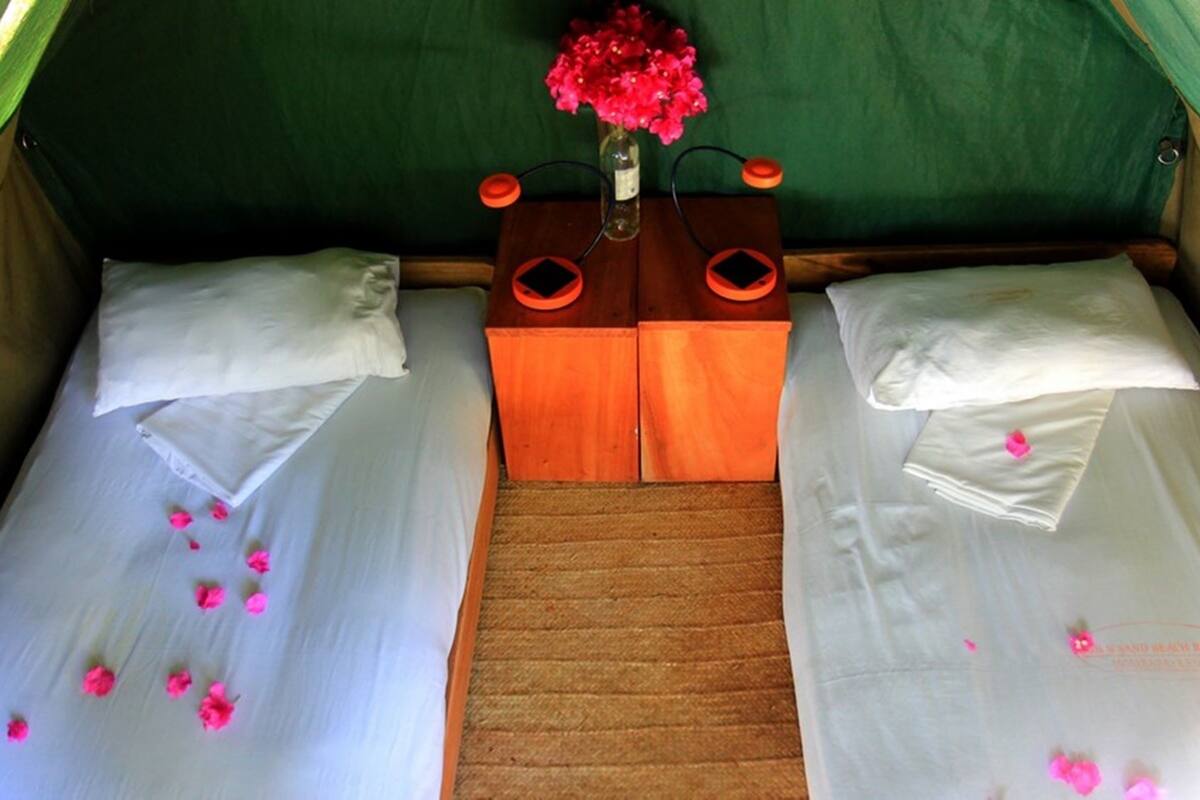 A tent-style room with two beds, each with white linens and pink flower petals scattered on them. A small wooden table with two orange lamps is placed between the beds.