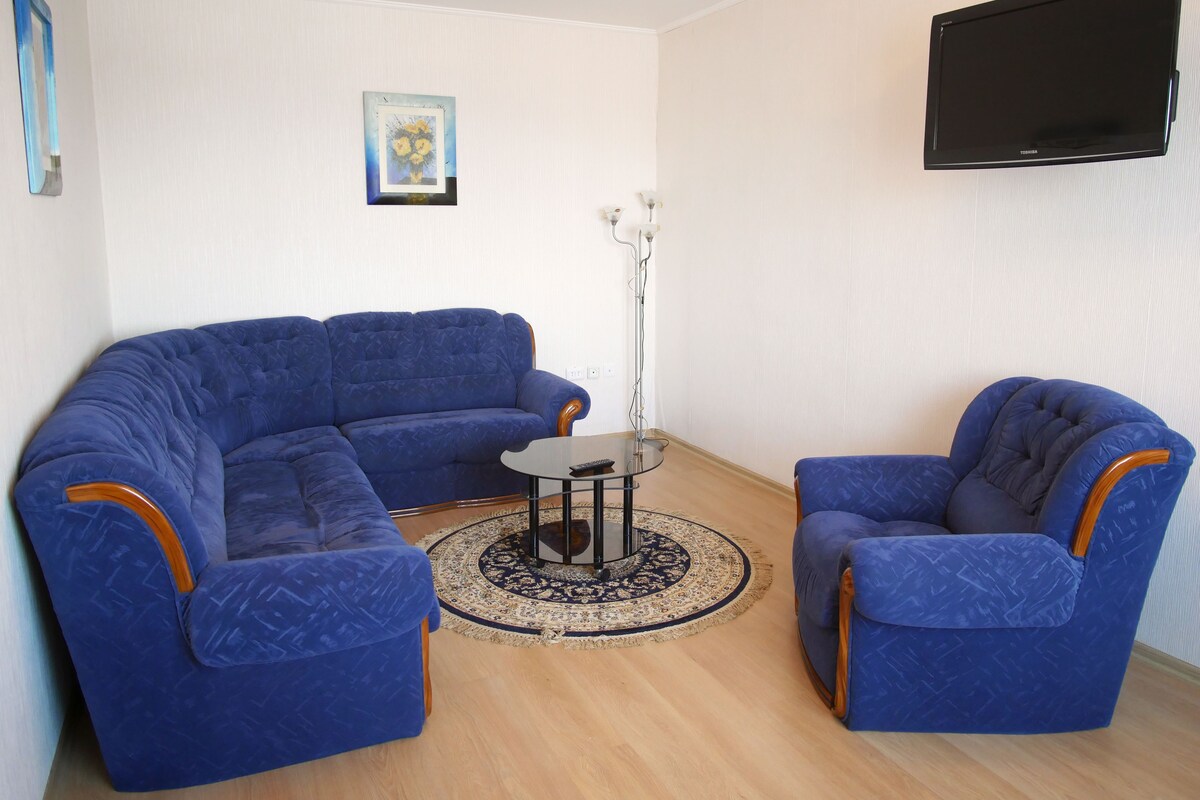 Un salon comprenant un canapé d’angle bleu, deux fauteuils assortis, une petite table basse, un lampadaire et un téléviseur à écran plat fixé au mur.