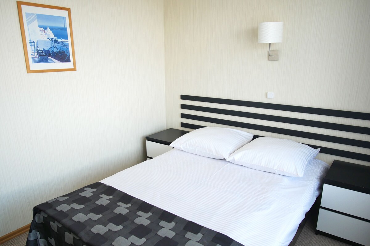 Une chambre d’hôtel comprenant un lit, une table de chevet, une commode, une lampe fixée au mur et un tableau encadré accroché au mur.
