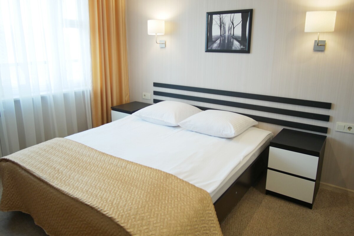 Une chambre d’hôtel comprenant un lit, deux tables de chevet, une commode, une fenêtre avec des rideaux et deux lampes fixées au mur.