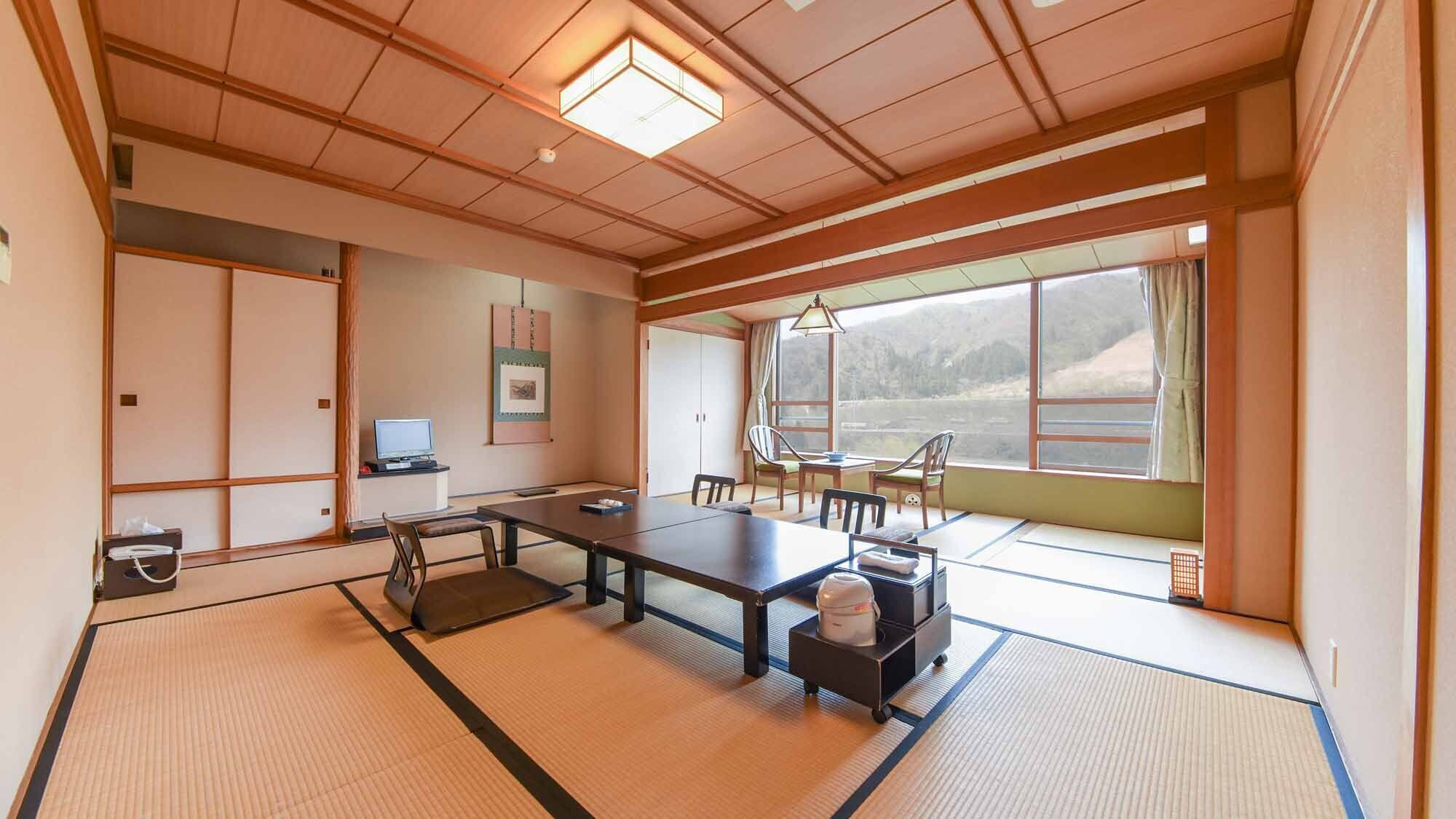 A traditional tatami mat room with a tatami mat table, chairs, a television, and a large window with a view of a mountainous landscape.