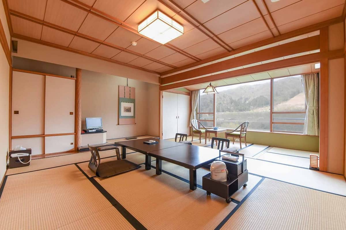 A traditional tatami mat room with a tatami mat table, chairs, a television, and a large window with a view of a mountainous landscape.