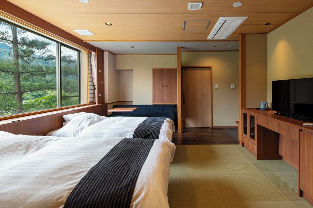 Habitación de hotel con dos camas, un escritorio con televisor y vistas a árboles a través de amplias ventanas.