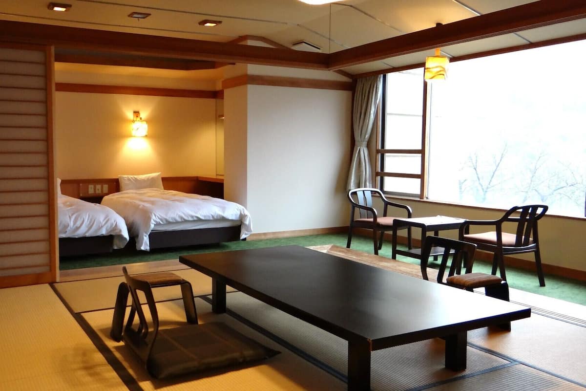 A hotel room with two beds, a desk, chairs, a table, and a view of trees through the window.