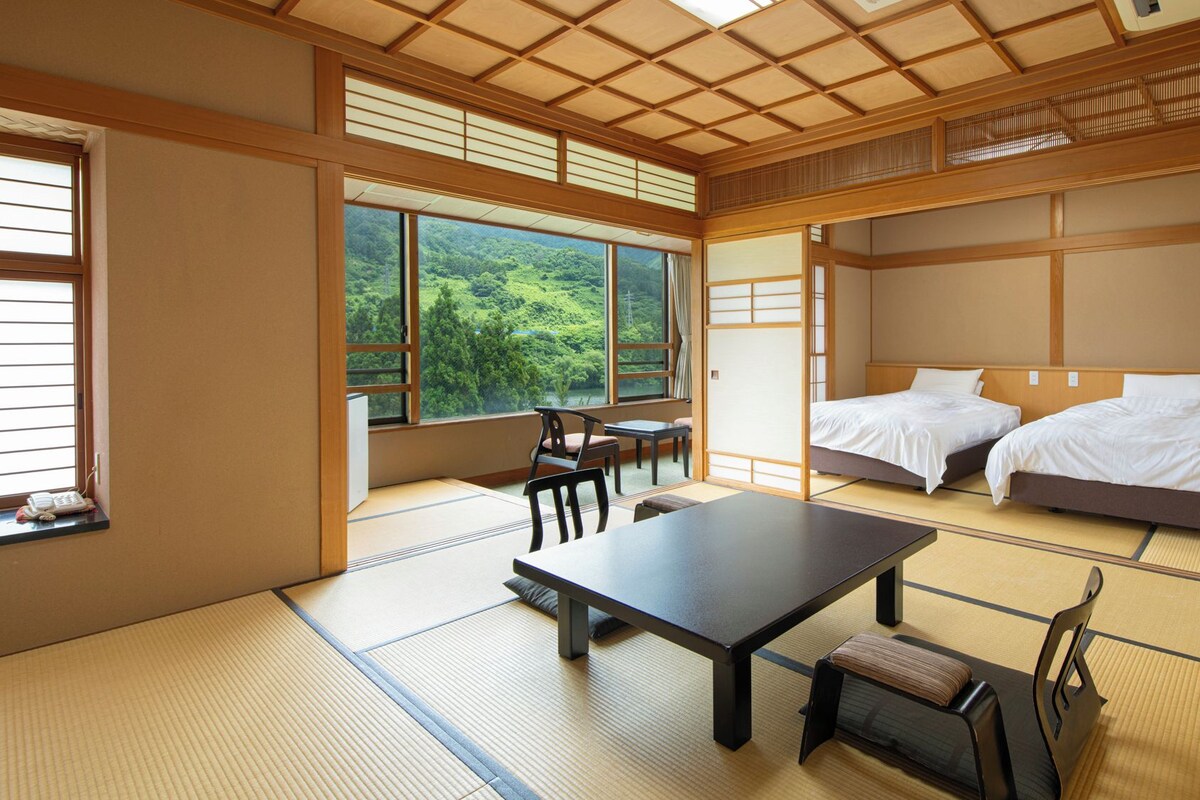 A traditional Japanese room with tatami flooring, a low table, two beds, and large windows offering a view of a forest.