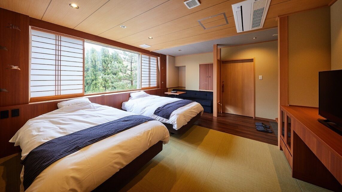 A hotel room with two beds, a TV, a desk, and a view of trees through the window.
