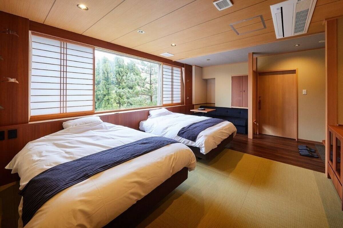 A hotel room with two beds, a TV, a desk, and a view of trees through the window.