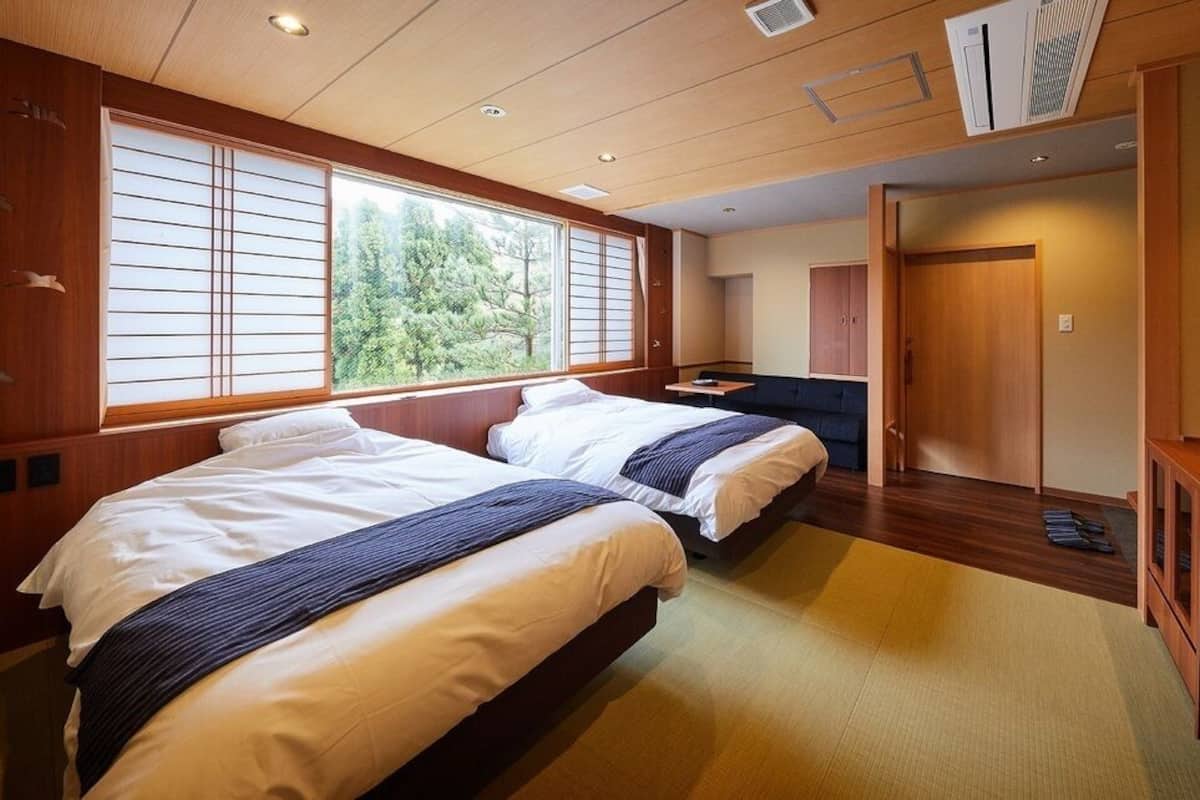 A hotel room with two beds, a TV, a desk, and a view of trees through the window.