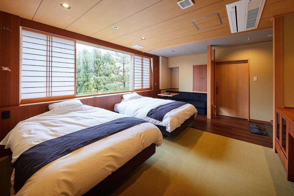 A hotel room with two beds, a TV, a desk, and a view of trees through the window.