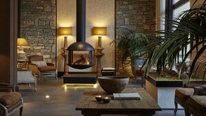 A cozy lounge area with stone walls, a fireplace, comfortable seating, and a large wooden table with a bowl and candles. - Limneon Resort & Spa (Kastoria)