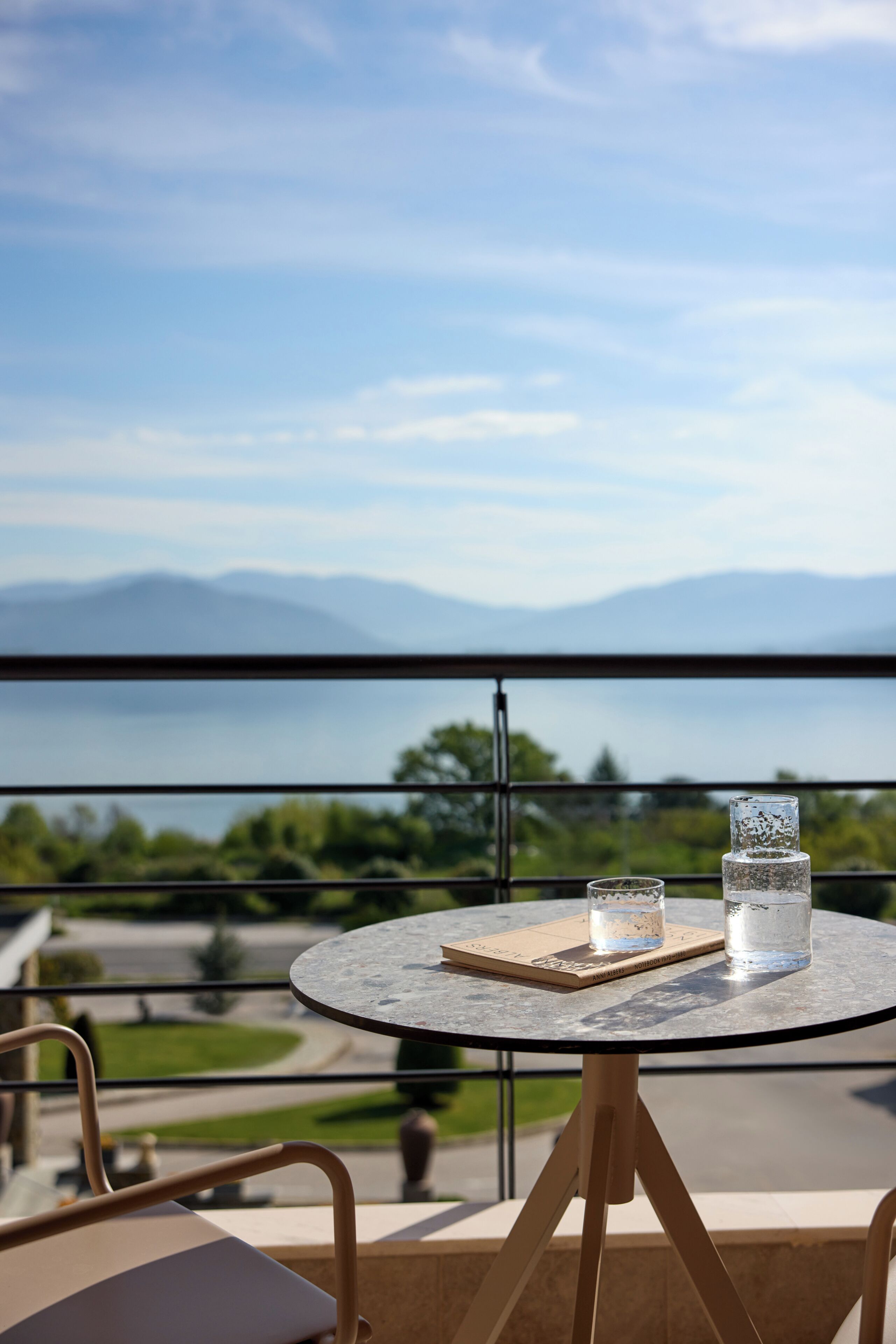 A round table with a glass and a book on a balcony overlooking a body of water and mountains.