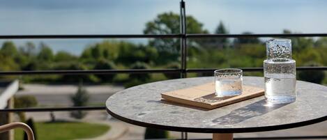 A round table with a glass and a book on a balcony overlooking a body of water and mountains.