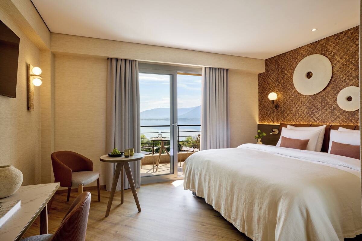 A hotel room with a bed, a desk, a chair, a small table with a plant, and a balcony with a view.