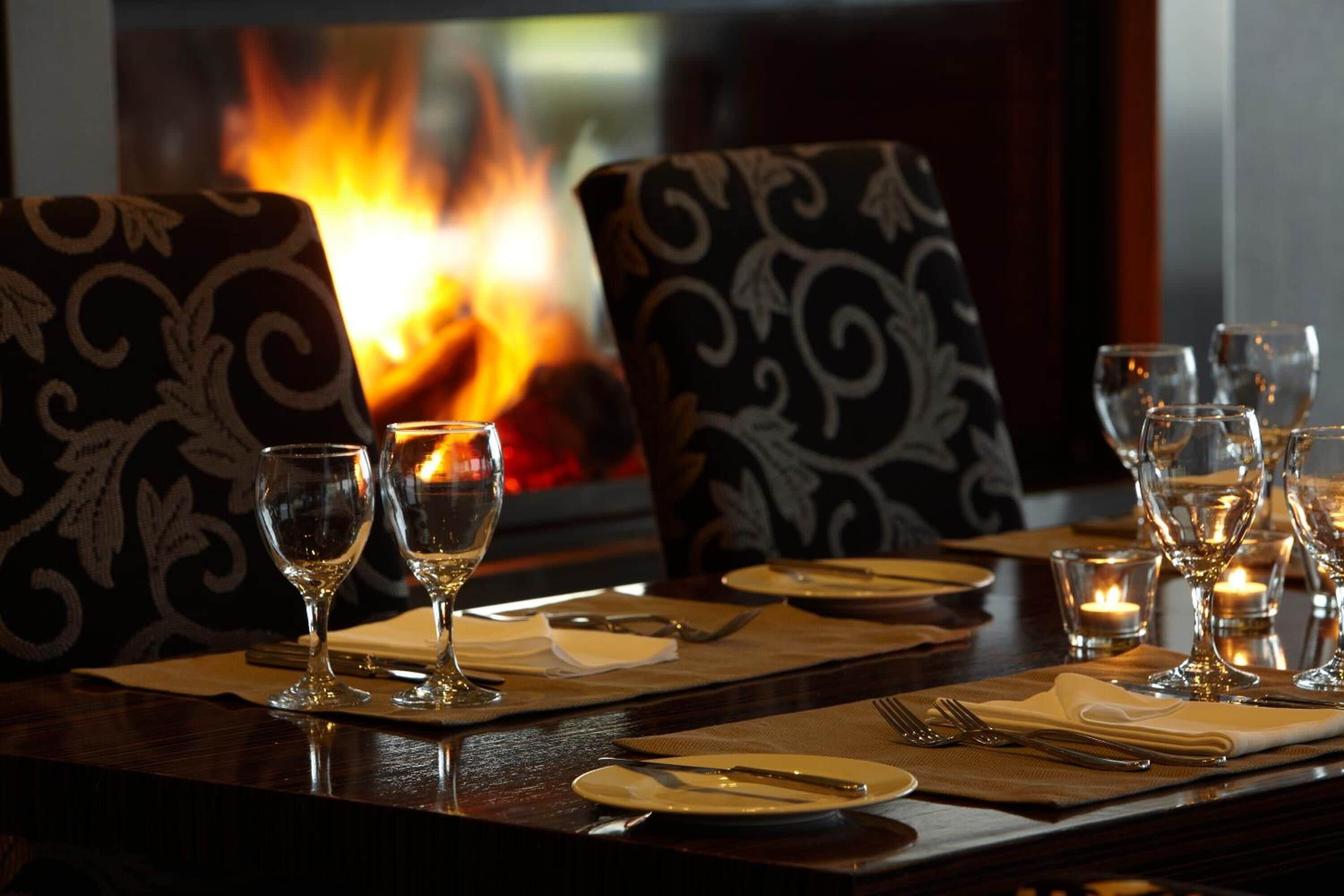 Een eettafel gedekt met wijnglazen, borden en bestek, met een open haard op de achtergrond.