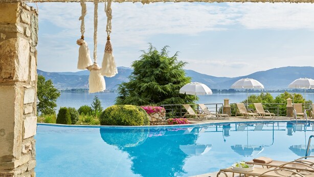 A pool area with a view of mountains and a lake, surrounded by lounge chairs and umbrellas. - Limneon Resort & Spa (Kastoria)