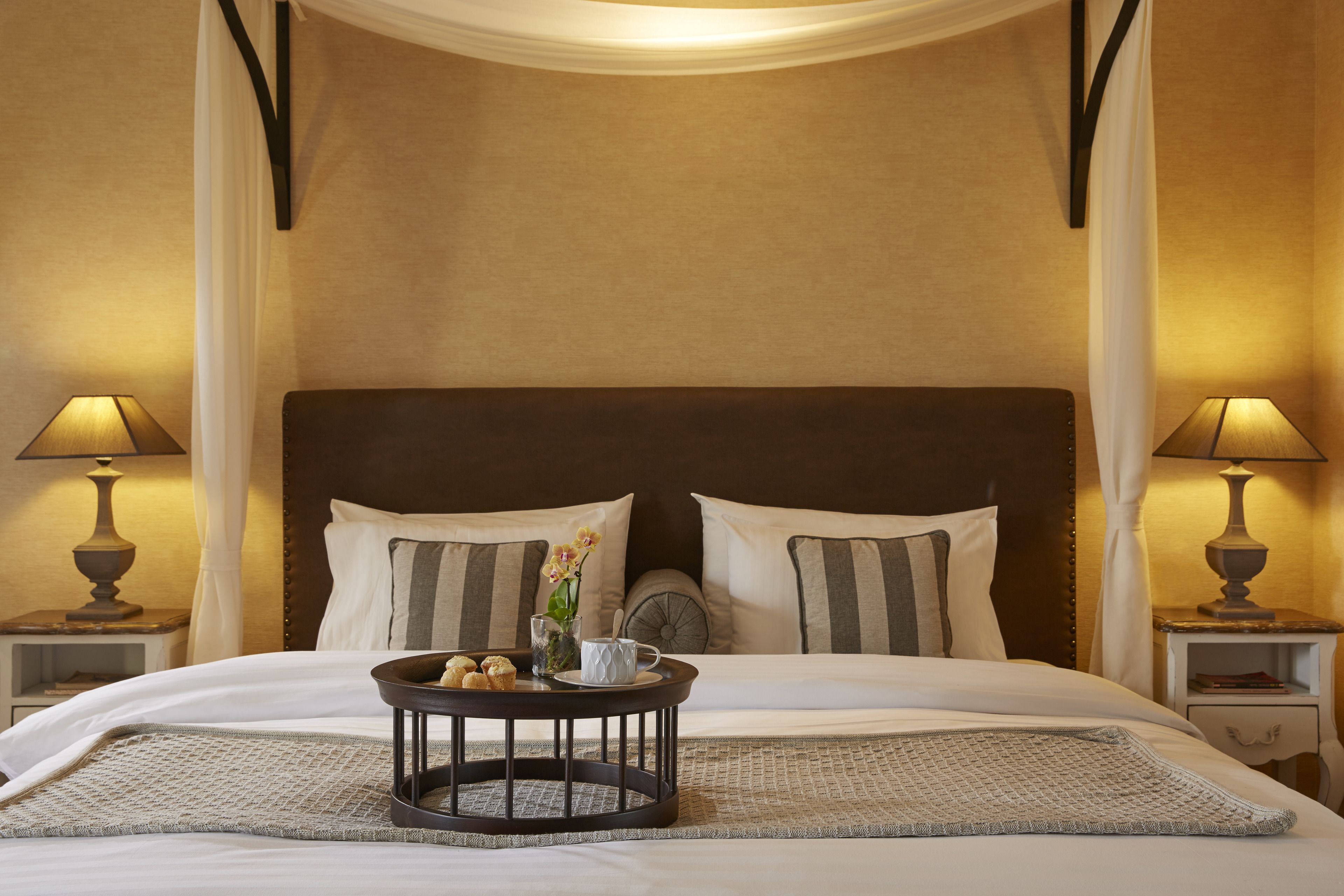 Una cama bien hecha con una mesita redonda, dos lámparas de noche y un jarrón decorativo con flores.