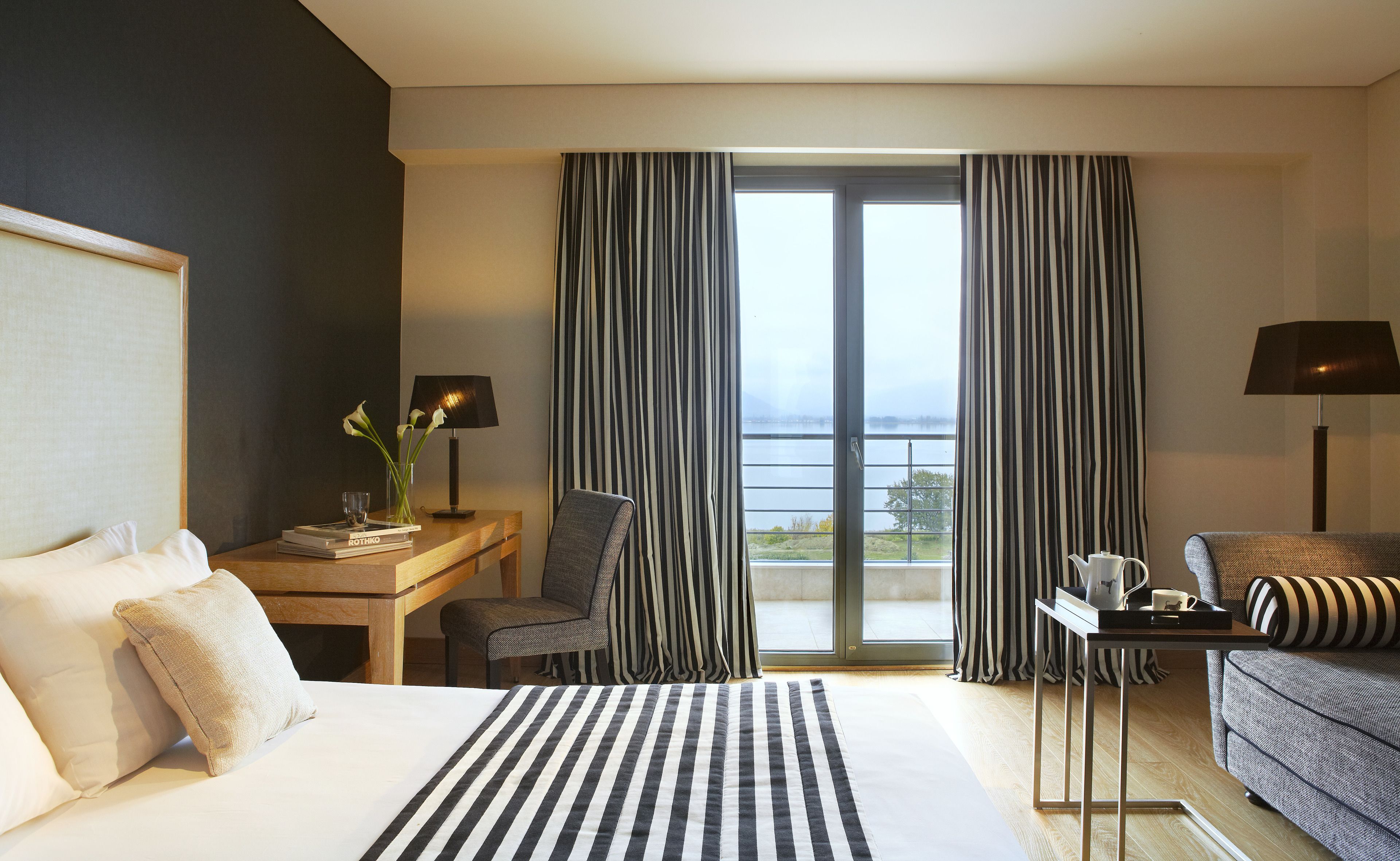 A hotel room with a bed, a desk with a lamp, a chair, a sofa, and a view of the outdoors.