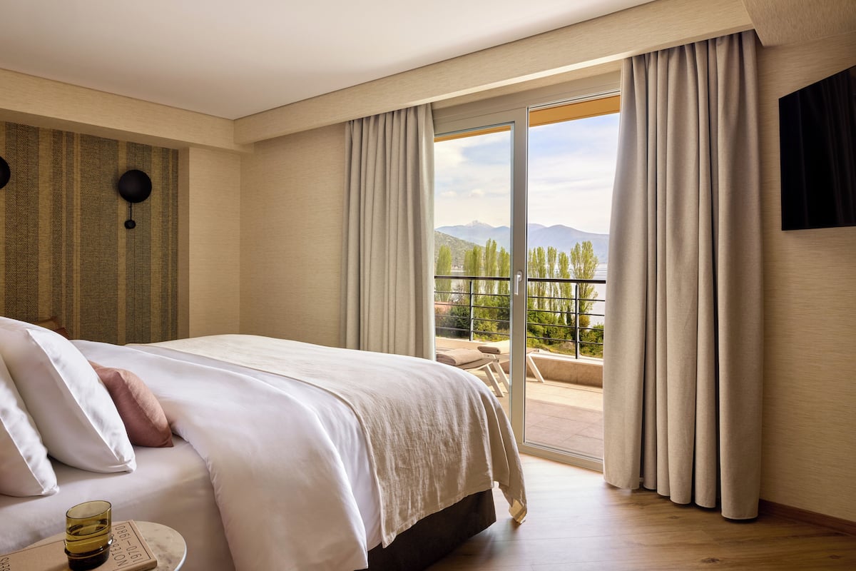 A hotel room with a bed, a view of the outdoors, a TV on the wall, and a window with curtains.