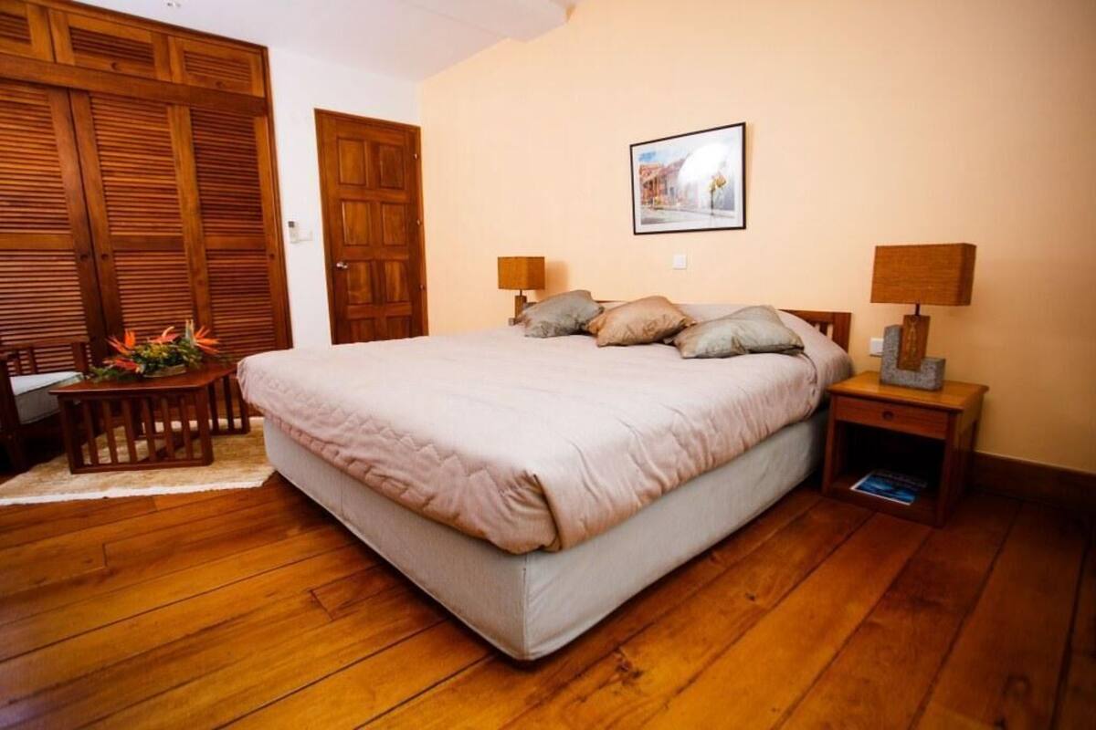 A bedroom with a large bed, wooden flooring, a small table with a lamp, and a picture on the wall.