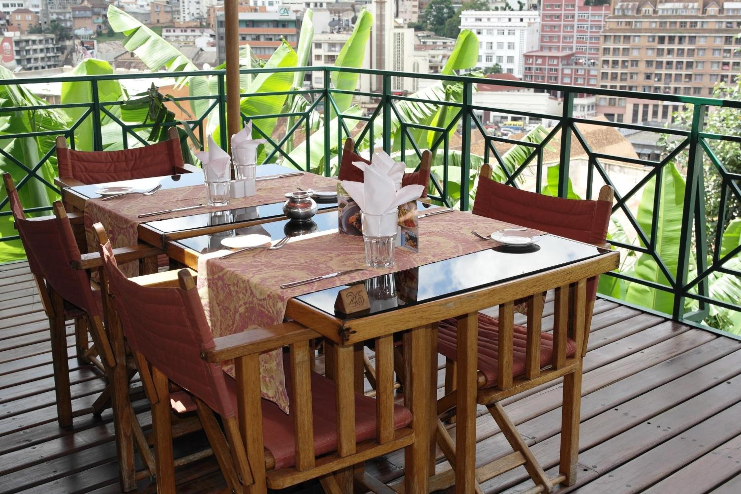 Un comedor con una mesa dispuesta para dos personas, rodeado de sillas de madera con cojines rojos, sobre una terraza de madera con vistas a edificios y follaje.