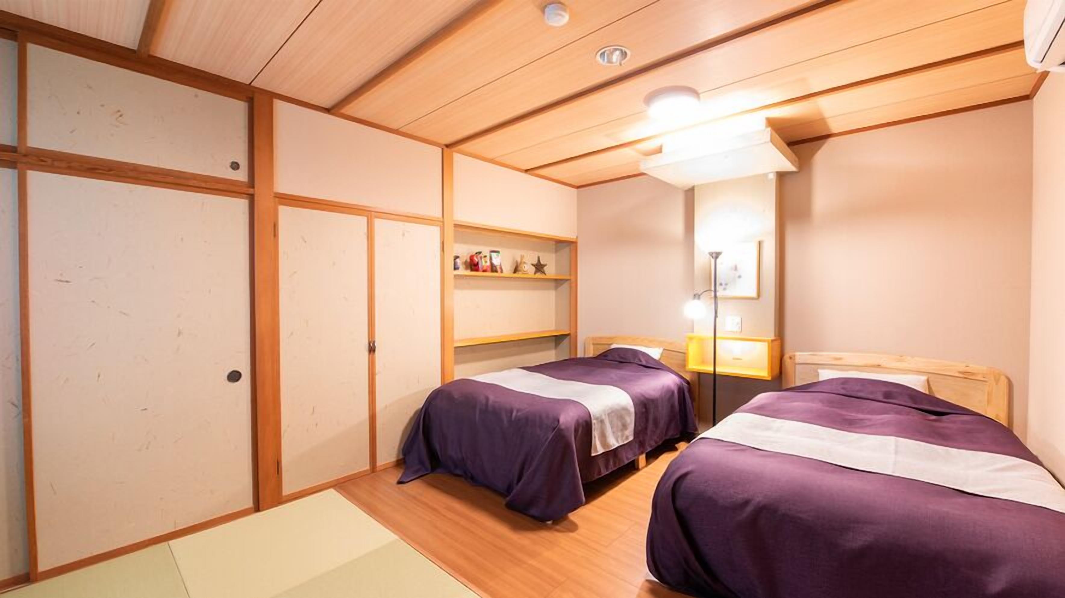 A room with two beds, wooden flooring, a shelf with items, and a mirror.
