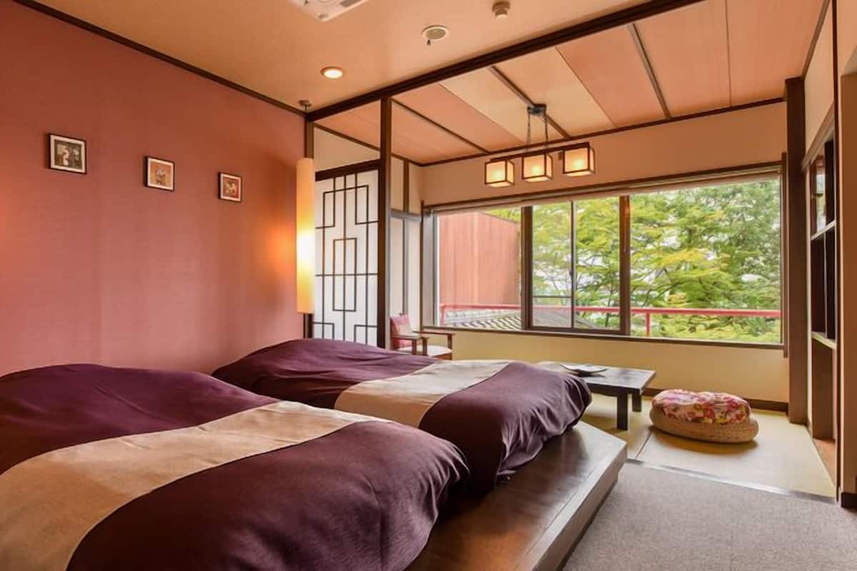 Une chambre d’hôtel avec deux lits, une petite table, une fenêtre donnant sur un paysage verdoyant et une porte coulissante de style japonais traditionnel.