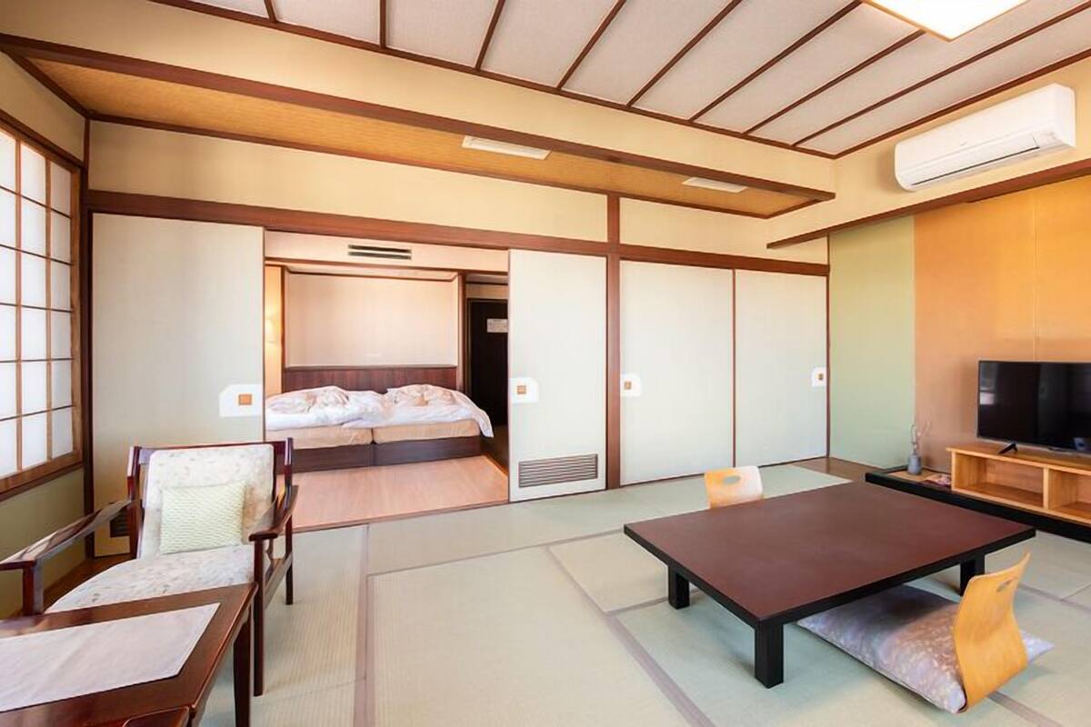 Une pièce de style japonais traditionnel, avec un sol recouvert de tatamis, une table basse, un canapé, un lit avec un futon et un téléviseur posé sur un meuble en bois.