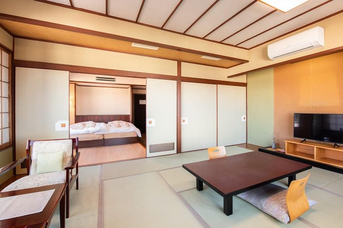 Une pièce de style japonais traditionnel, avec un sol recouvert de tatamis, une table basse, un canapé, un lit avec un futon et un téléviseur posé sur un meuble en bois.