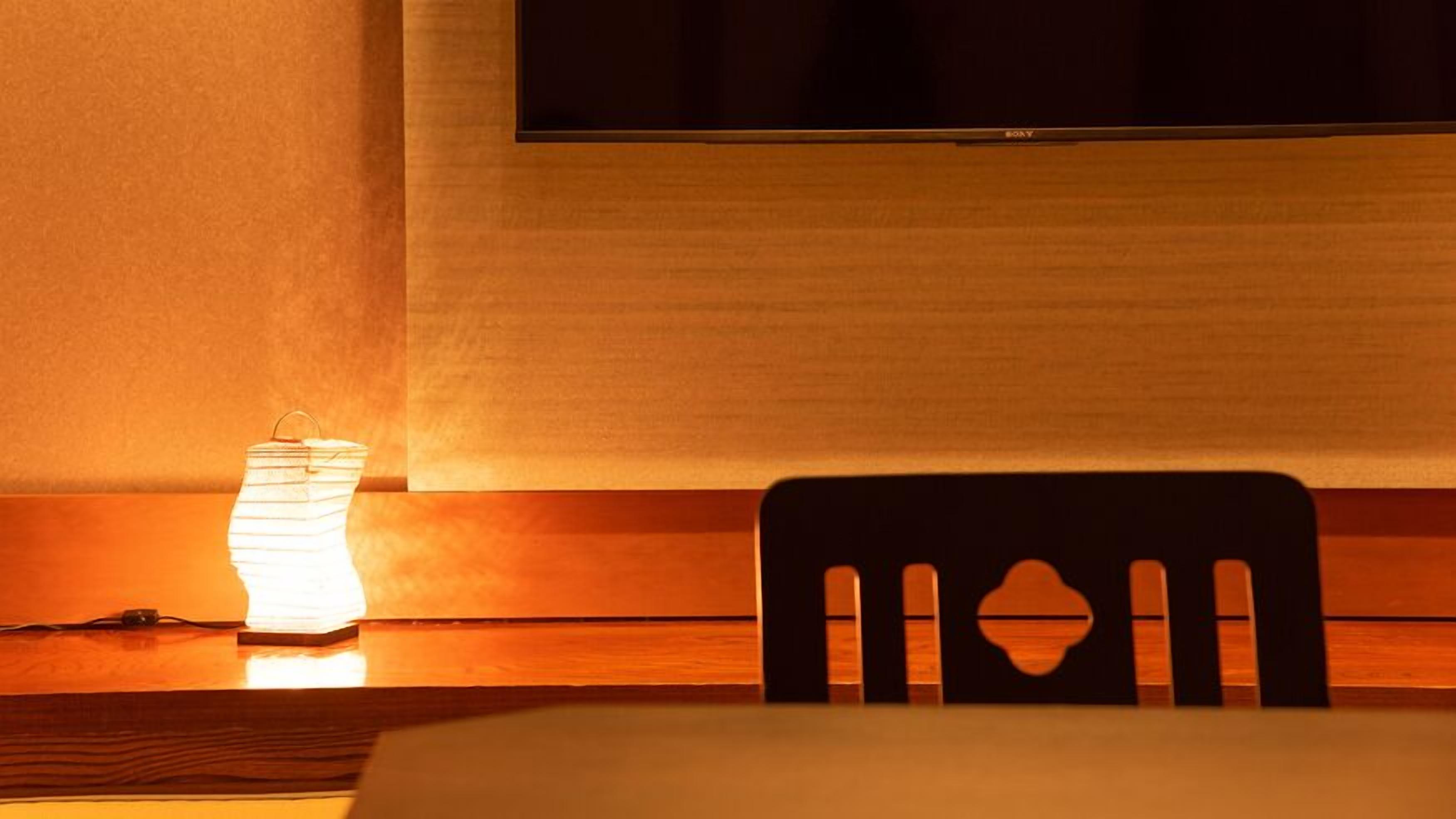A hotel room with a television mounted on the wall, a wooden chair, and a table with a lamp.