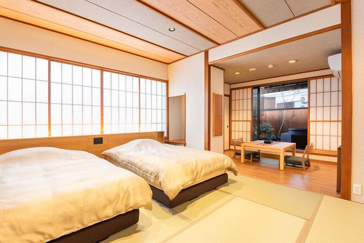 A traditional Japanese-style room with tatami flooring, two beds, a sliding door leading to a balcony, and a low wooden ceiling.