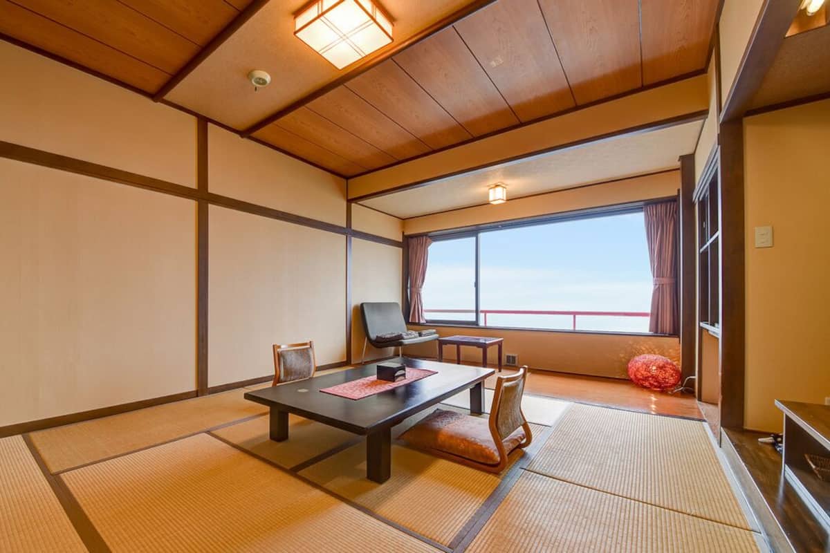 Une pièce de style japonais traditionnel, avec un sol recouvert de tatamis, une table basse et une vue dégagée sur l’extérieur grâce à une grande fenêtre.