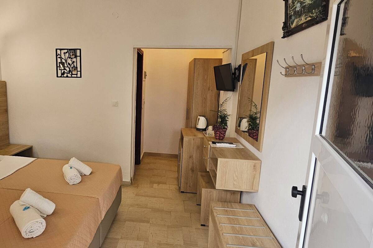 Quarto de hotel com cama, duas mesas de cabeceira, painel de cama de madeira, pequena escrivaninha com espelho e um cabideiro.