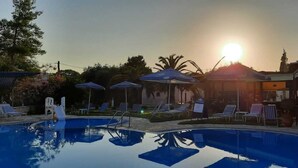 A swimming pool with lounge chairs and umbrellas, surrounded by buildings and trees. - Pension Nychterida (Corfu)
