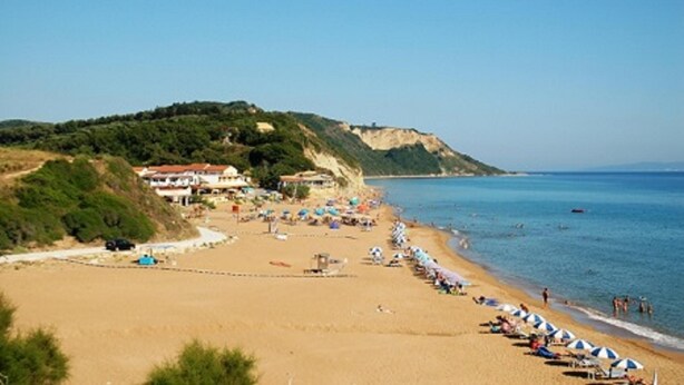 A beach with rows of umbrellas and beach chairs, a building with a red roof, and a cliff in the background. - Pension Nychterida (Corfu)