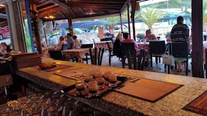 An outdoor restaurant with tables and chairs, a counter with glasses and a tray, and a view of the street. - Pension Nychterida (Corfu)